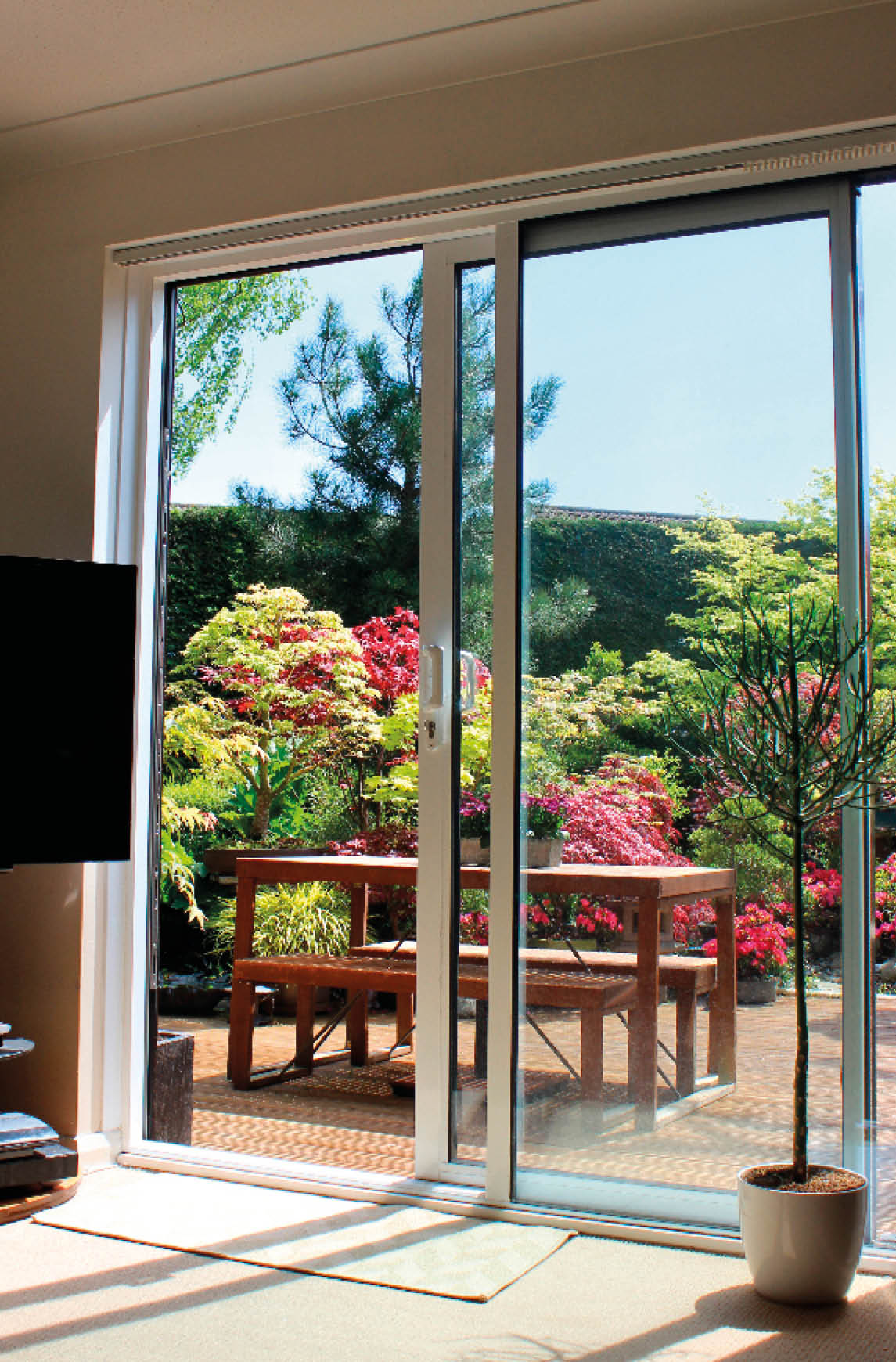 Photo showing some modern aluminium patio doors that are part of a lounge / sitting room and positioned next to the flat-screen television. The patio doors are pictured partly open, serving as a window and doorway into the beautifully landscaped garden and its timber decking area, helping to 'bring the outside in'.